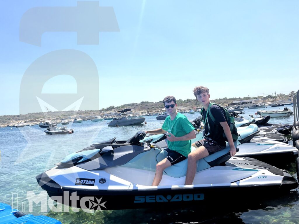 Two students smile from a jet ski moored in a marina, with calm blue water and boats behind them.