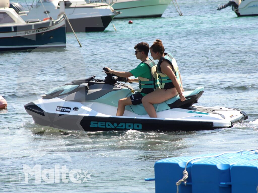 Two young participants ride a Sea-Doo through a marina area, wearing life jackets with moored boats and buoys nearby.