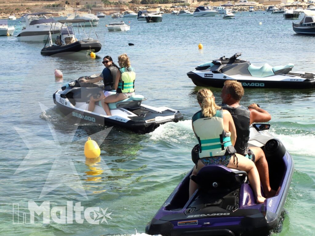 Two pairs of young participants ride jet skis through a busy Mediterranean bay, leaving wakes near boats and colorful buoys.