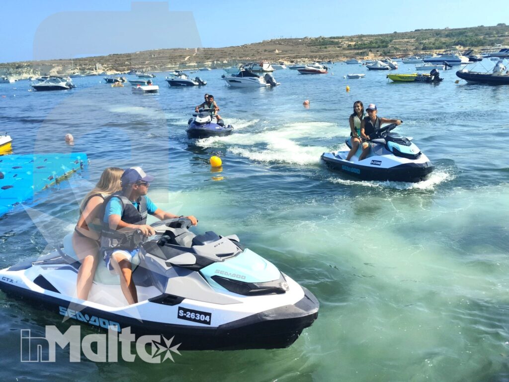 Two people ride a jet ski through a crowded Mediterranean bay, with spray, turquoise water, boats, and other participants around them.