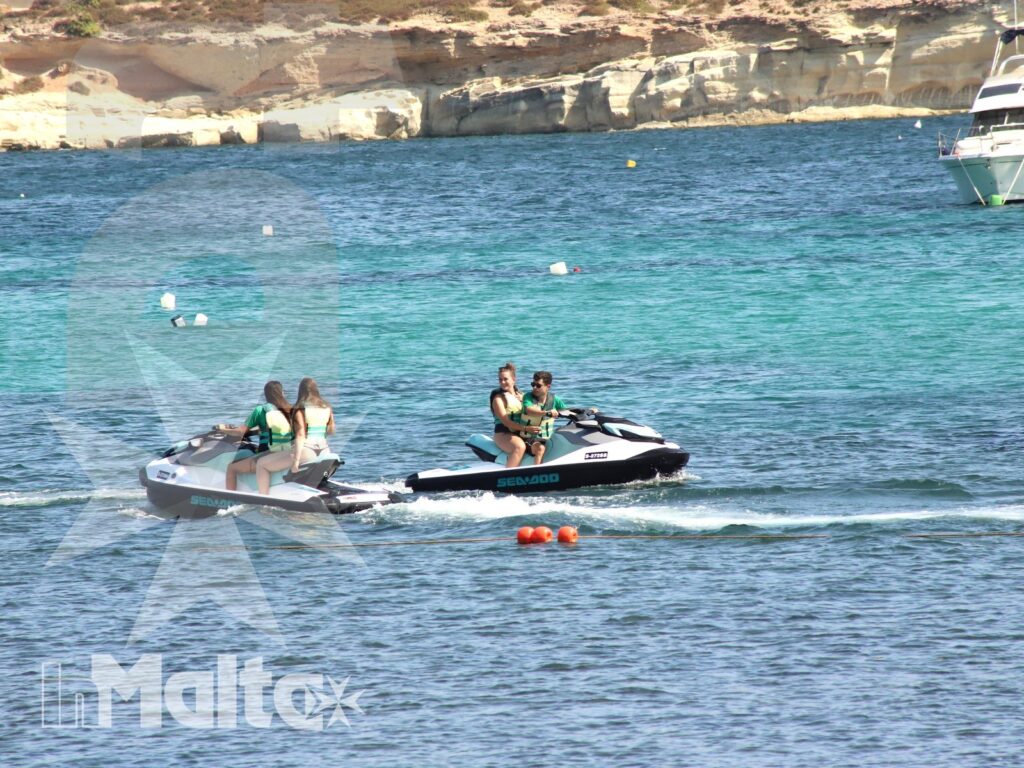 Two jet skis cross a turquoise Mediterranean bay, their parallel wakes contrasting with pale cliffs and anchored boats.