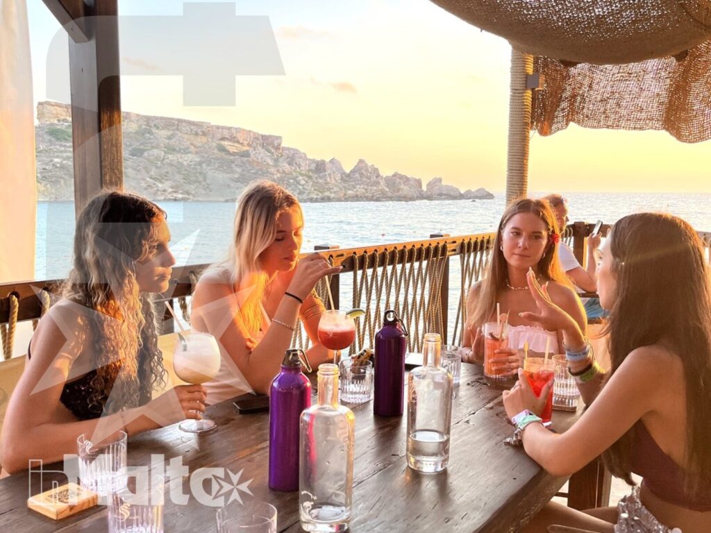 Young women toasting during an inMalta coastal experience Four young women raise colorful cocktails around an outdoor table, enjoying a social seaside experience with inMalta.