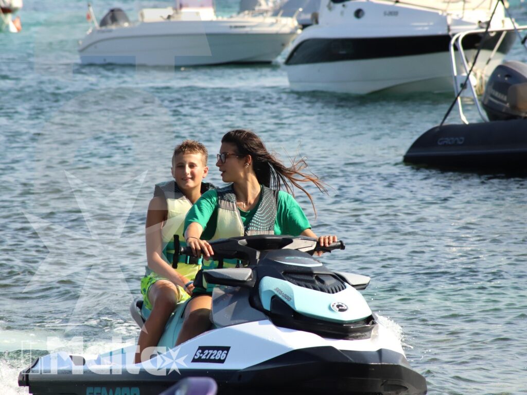 Two young participants ride a jet ski together, wearing life jackets as motorboats and reflections fill the coastal water behind them.