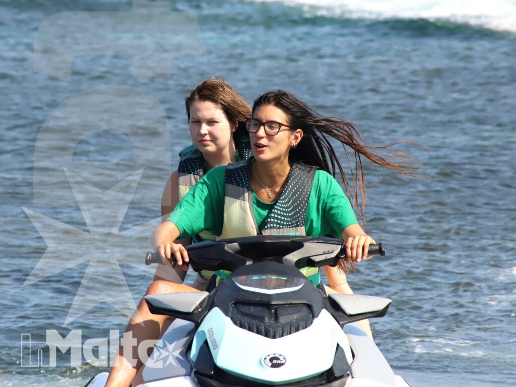 Two young women navigate a jet ski across open water, with windblown hair, small waves, and spray showing the movement.