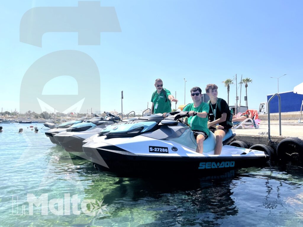 Three students sit on a jet ski at a marina while an instructor stands behind them during supervised preparation.