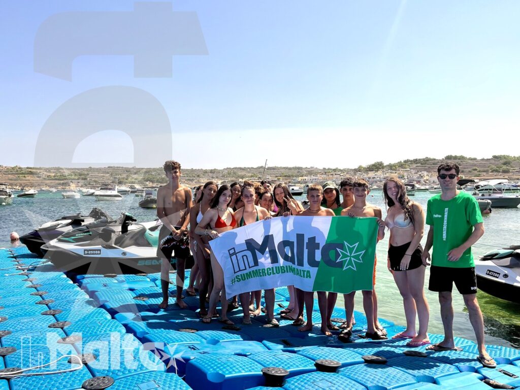 Students gather on a floating dock beside jet skis while holding a school sign during an organized water activity in Malta.
