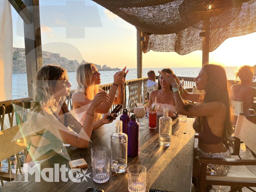 Participants toasting during an inMalta seaside excursion A group of young adults toast and chat around a table on a seaside terrace during an inMalta boat excursion at sunset.