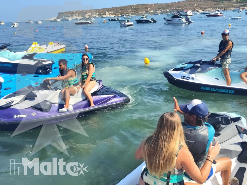 Young participants ride colorful jet skis through a clear Mediterranean bay, surrounded by anchored boats, buoys, and rocky coastline.