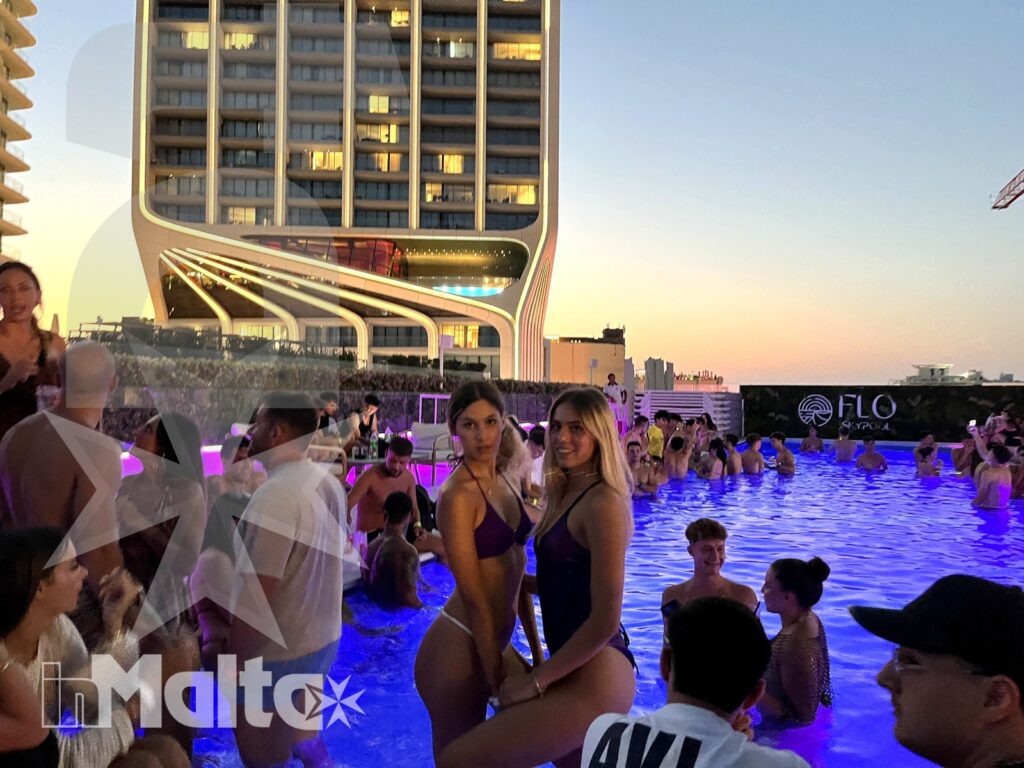 Two young women pose beside a crowded rooftop pool at sunset as guests swim and socialize beneath blue and purple lights.