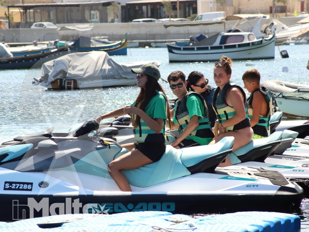 A group of young people in life jackets sits on stationary jet skis in a sunny marina surrounded by boats.