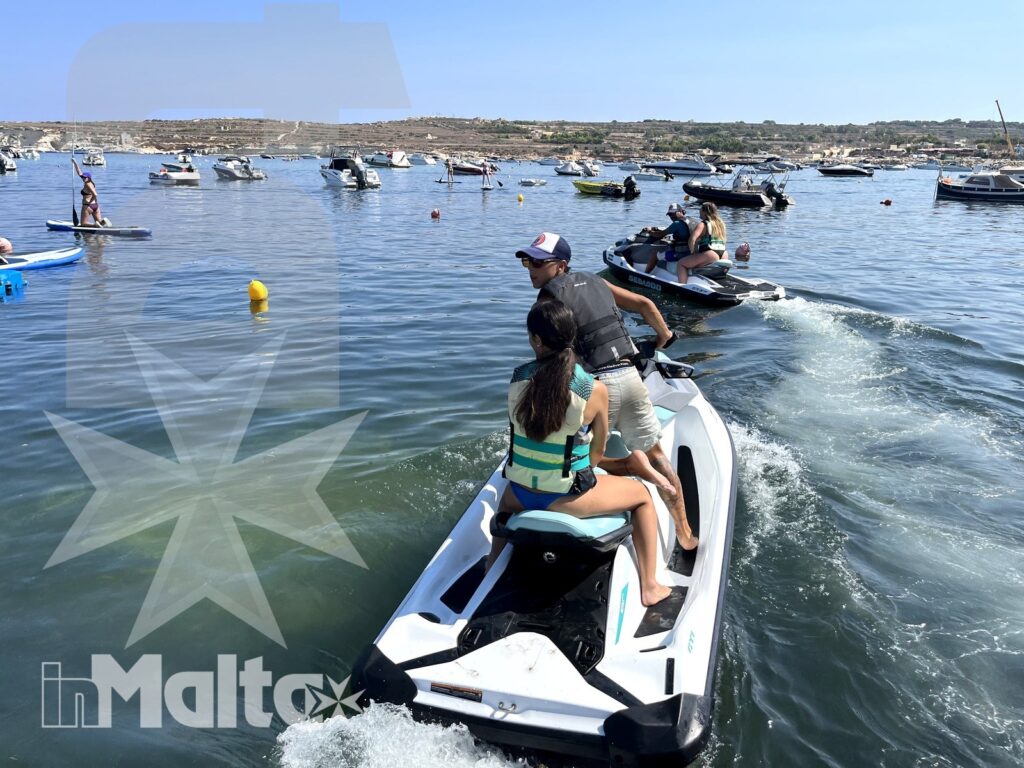 Two young participants navigate a jet ski through a marina, leaving a wake among colorful buoys, boats, and coastal scenery.