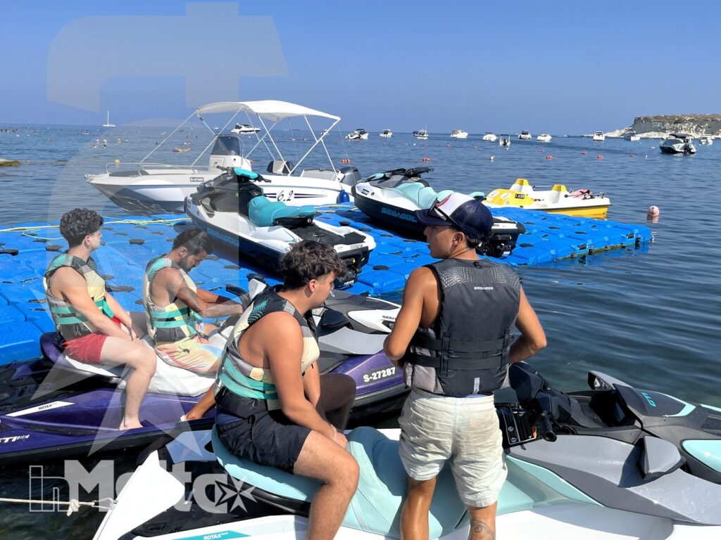 Young participants and an adult organize jet skis and safety equipment on a floating dock in a Mediterranean bay.