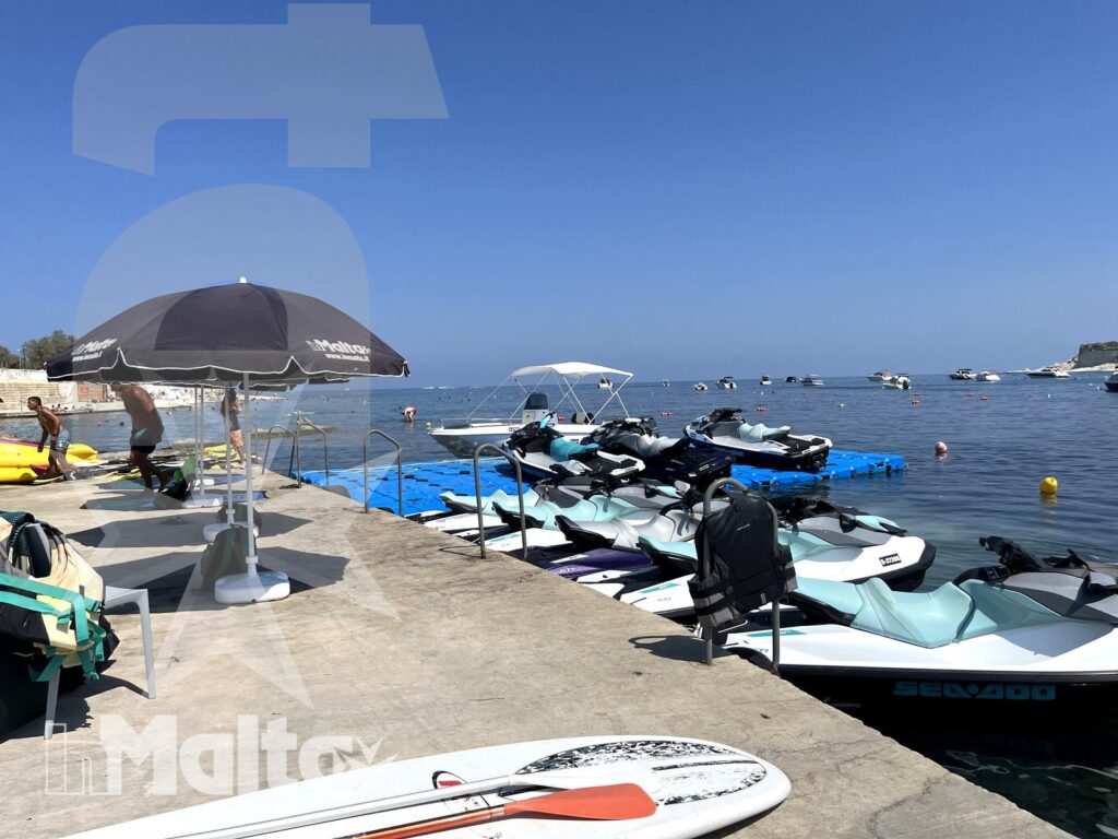 A marina launch area displays parked jet skis, paddle boards, a floating platform, and umbrellas beside calm open water.