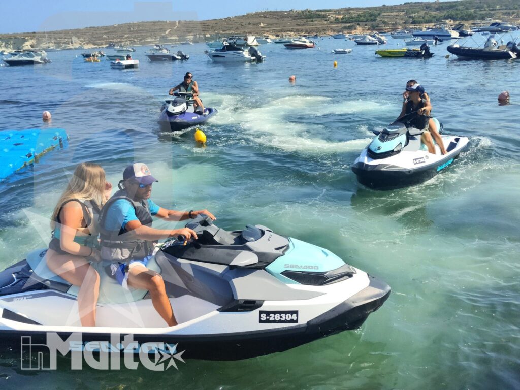 Participants steer three jet skis across a sunny Mediterranean bay, leaving visible wakes among anchored boats and rocky shores.