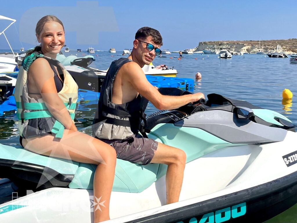 Two smiling students sit on a turquoise and white jet ski, with boats, buoys, and rocky coastline visible behind them.