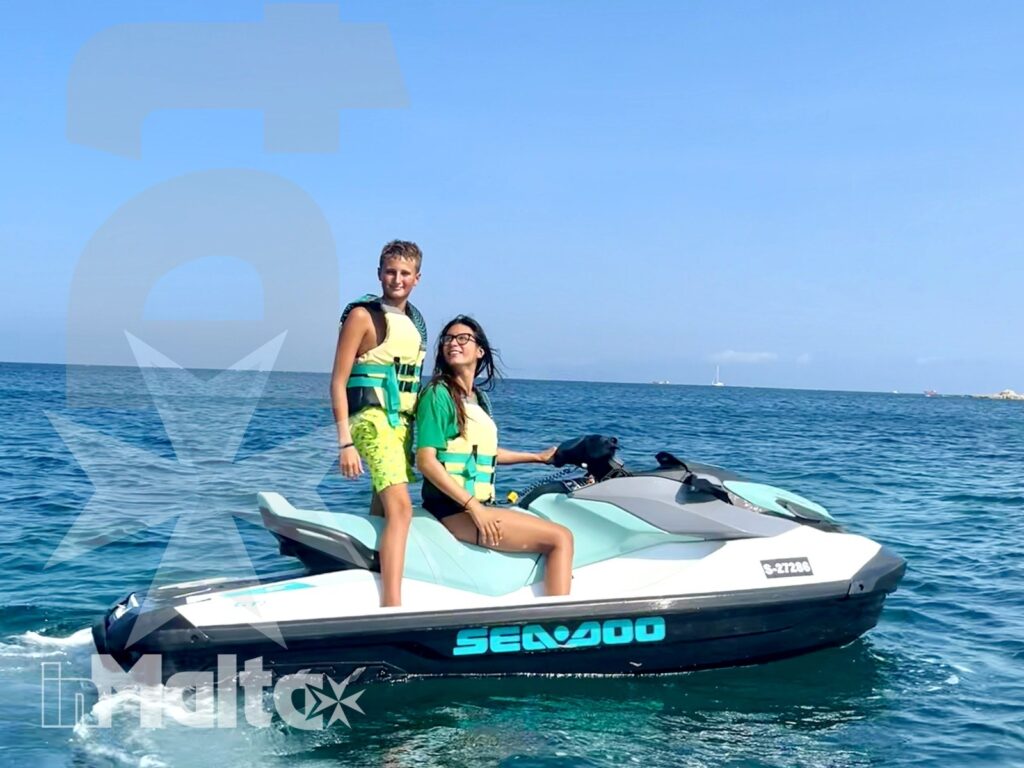 Two young people wearing life jackets pose together on a jet ski in calm open sea beneath a bright blue sky.